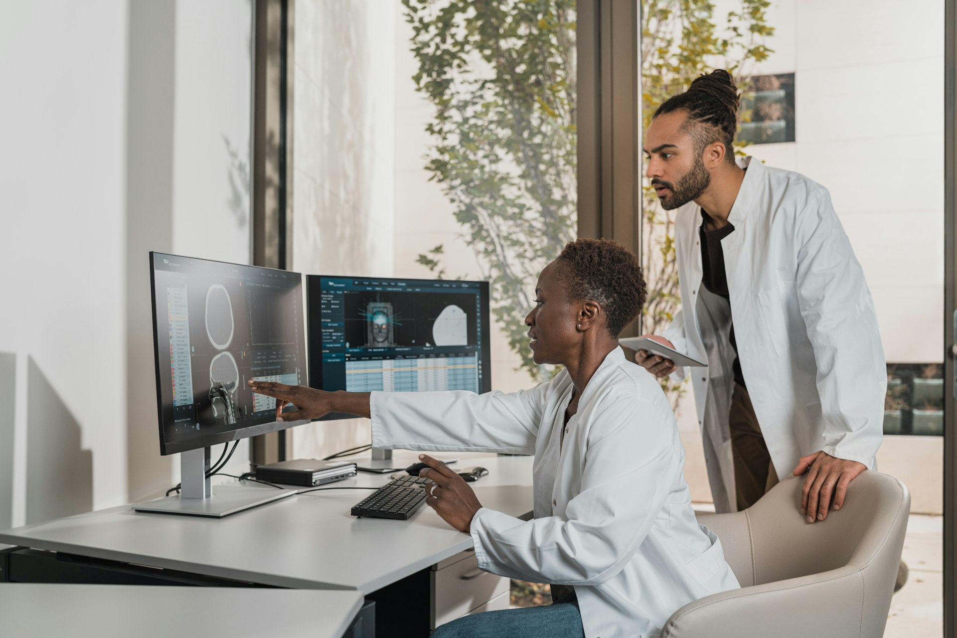 A clinician in a white coat reviews scans and a data table on two monitors while a colleague looks on.